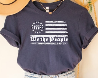 May include: A navy blue t-shirt with a white distressed American flag design and the text "1776 We the People".
