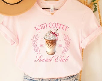 May include: A pink V-neck t-shirt with the words "Iced Coffee Social Club" and an illustration of an iced coffee drink. The shirt is paired with denim shorts, a straw hat, and a woven crossbody bag.