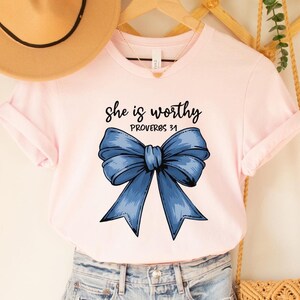 May include: Pink t-shirt with a blue bow and the text "She is worthy Proverbs 31" printed on it.