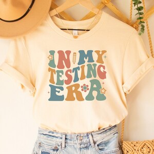 May include: A soft cream colored t-shirt with the text "In My Testing Era" in a retro font with colorful accents. The shirt is hanging on a wooden hanger with a straw hat and a woven bag.