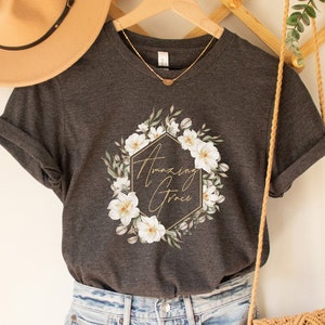 May include: Dark grey heather t-shirt with a floral wreath design and the text "Amazing Grace" in gold lettering.