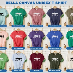 May include: A collection of Bella Canvas unisex t-shirts in various colors featuring a white silhouette of a fish with the word "MEOW" printed below. The colors include Heather Ice Blue, Heather Kelly, Heather Mauve, Heather Navy, Heather Olive, Heather Peach, Heather Red, Heather Team Purple, Heather True Royal, Kelly, Leaf, Light Blue, Maroon, Military Green, and Natural.
