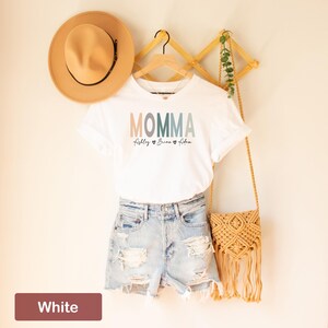 May include: White t-shirt with the word "MOMMA" in colorful letters and the names "Abigale", "Brian", and "Eden" written below. The shirt is hanging on a wooden hanger with a straw hat and a woven bag.