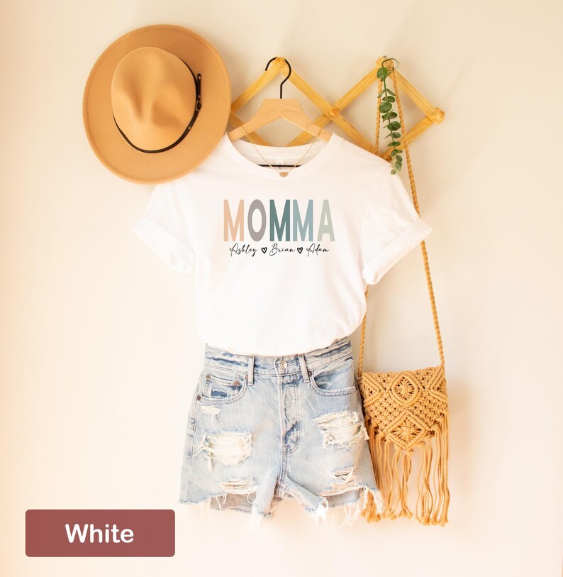 May include: White t-shirt with the word "MOMMA" in colorful letters and the names "Abigale", "Brian", and "Eden" written below. The shirt is hanging on a wooden hanger with a straw hat and a woven bag.