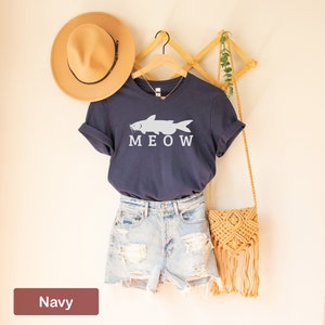 May include: A navy blue t-shirt with a white catfish graphic and the word "MEOW" printed below it. The shirt is hanging on a wooden hanger with a straw hat and a woven bag.
