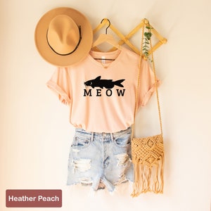May include: A peach colored t-shirt with a black catfish graphic and the text "MEOW". The shirt is hanging on a wooden hanger with a straw hat and a woven bag.