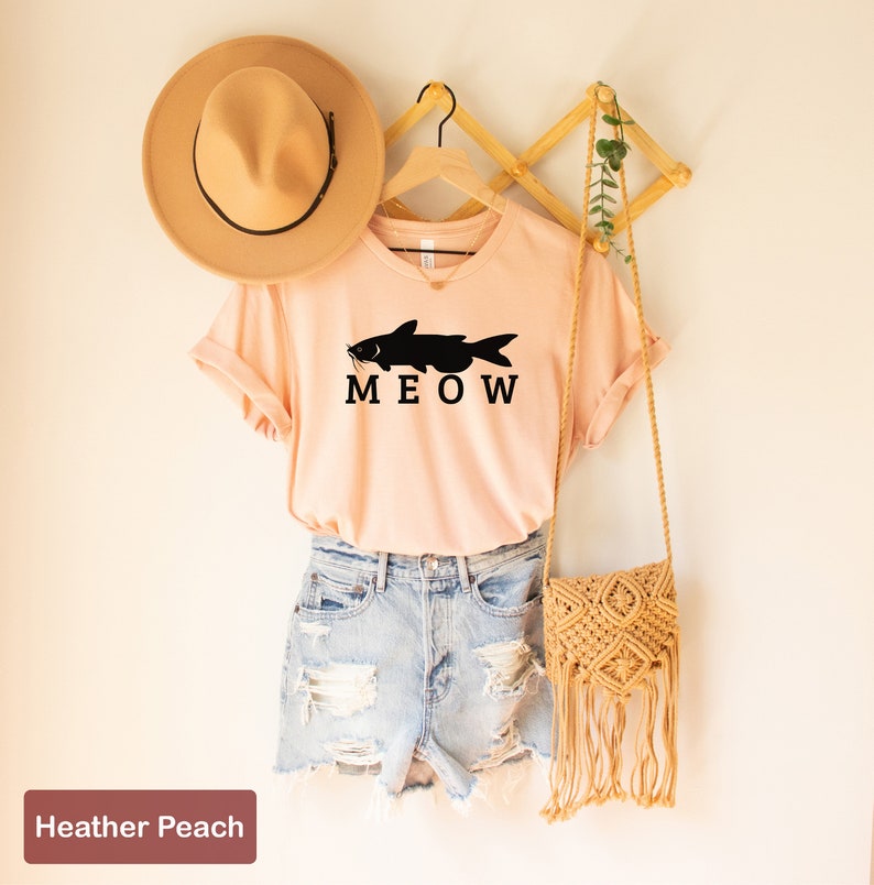 May include: A peach colored t-shirt with a black catfish graphic and the text "MEOW". The shirt is hanging on a wooden hanger with a straw hat and a woven bag.