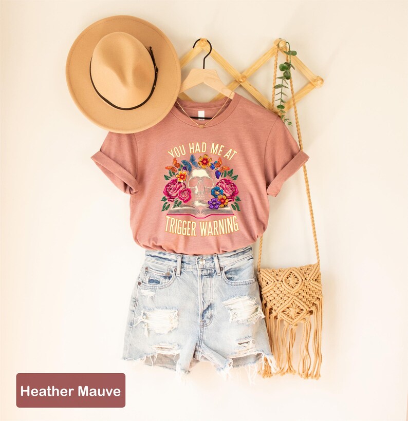 Puede incluir: Una camiseta de color malva jaspeado con el texto "You had me at trigger warning" y una ilustraci&oacute;n floral de una calavera y un libro. La camiseta est&aacute; modelada en una persona que lleva pantalones cortos de mezclilla y un sombrero de paja.
