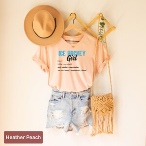 Op de afbeelding: Een perzikkleurig T-shirt met de tekst "ICE HOCKEY Girl (noun) 1. Like a normal girl only cooler... way cooler. see also: "sassy", "empowered", "fierce" erop gedrukt. Het shirt hangt aan een houten hanger met een bruine hoed en een geweven tas.