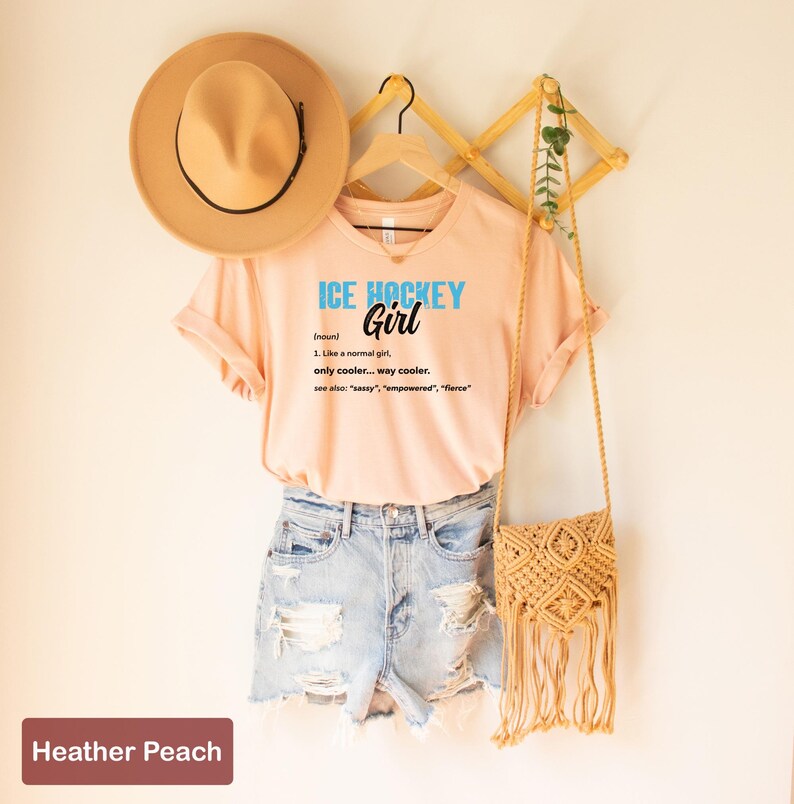 Op de afbeelding: Een perzikkleurig T-shirt met de tekst "ICE HOCKEY Girl (noun) 1. Like a normal girl only cooler... way cooler. see also: "sassy", "empowered", "fierce" erop gedrukt. Het shirt hangt aan een houten hanger met een bruine hoed en een geweven tas.