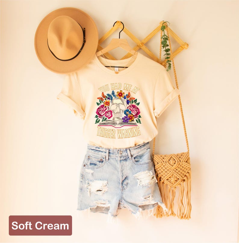 Puede incluir: Una camiseta de color crema con un gr&aacute;fico de calavera y flores y el texto "You had me at trigger warning".