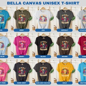 Puede incluir: Una maqueta de una camiseta unisex en 15 colores diferentes con una calavera con flores y el texto "You Had Me At Trigger Warning".