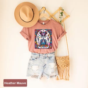 May include: A heather mauve t-shirt featuring a colourful graphic of a skeleton holding a paintbrush and the text "The Crafter". The shirt is styled with denim shorts and a straw bag.