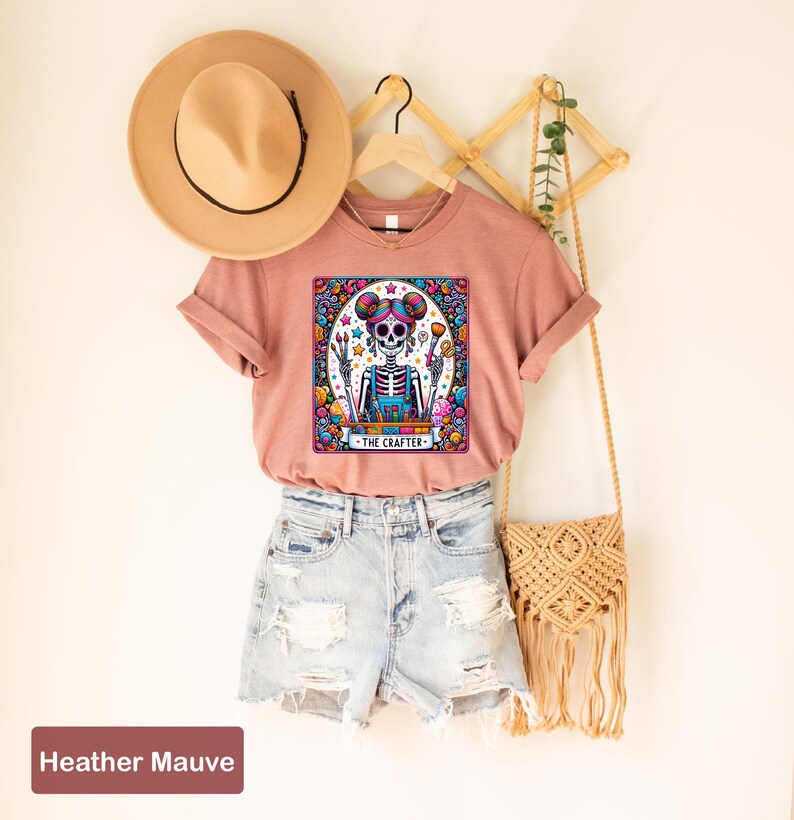 May include: A heather mauve t-shirt featuring a colourful graphic of a skeleton holding a paintbrush and the text "The Crafter". The shirt is styled with denim shorts and a straw bag.