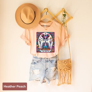 May include: A peach coloured t-shirt featuring a colourful graphic of a skeleton holding paintbrushes and a paint palette. The graphic is surrounded by a colourful border and the text "The Crafter" is below the skeleton.