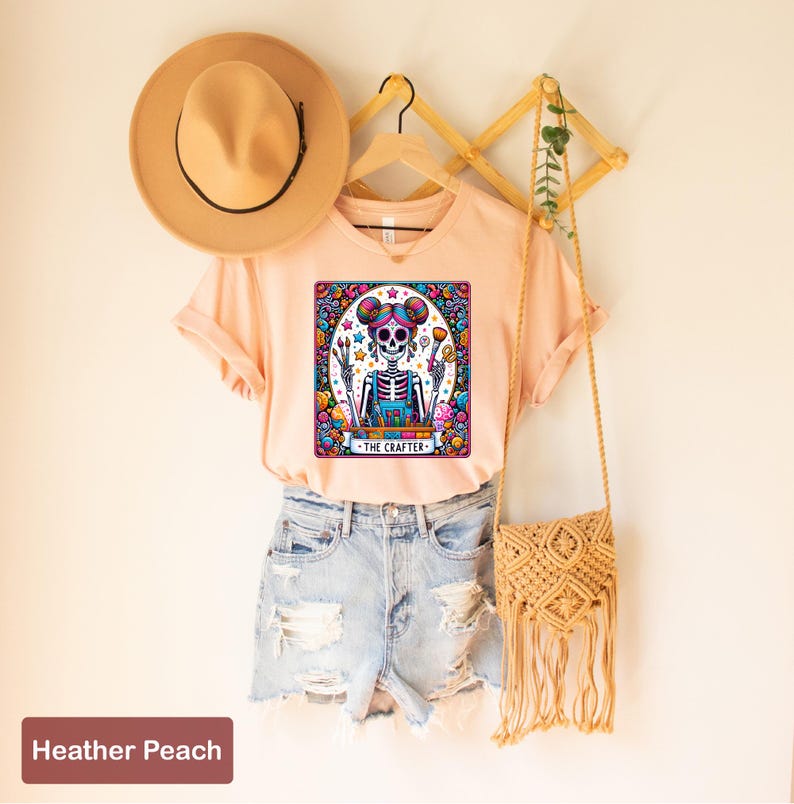 May include: A peach coloured t-shirt featuring a colourful graphic of a skeleton holding paintbrushes and a paint palette. The graphic is surrounded by a colourful border and the text "The Crafter" is below the skeleton.