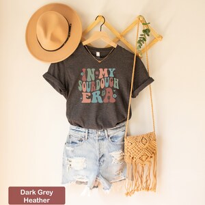 May include: Dark grey heather t-shirt with a retro-style graphic that says "In My Sourdough Era" with flowers and hearts. The shirt is hanging on a wooden hanger with a straw hat and a woven bag.