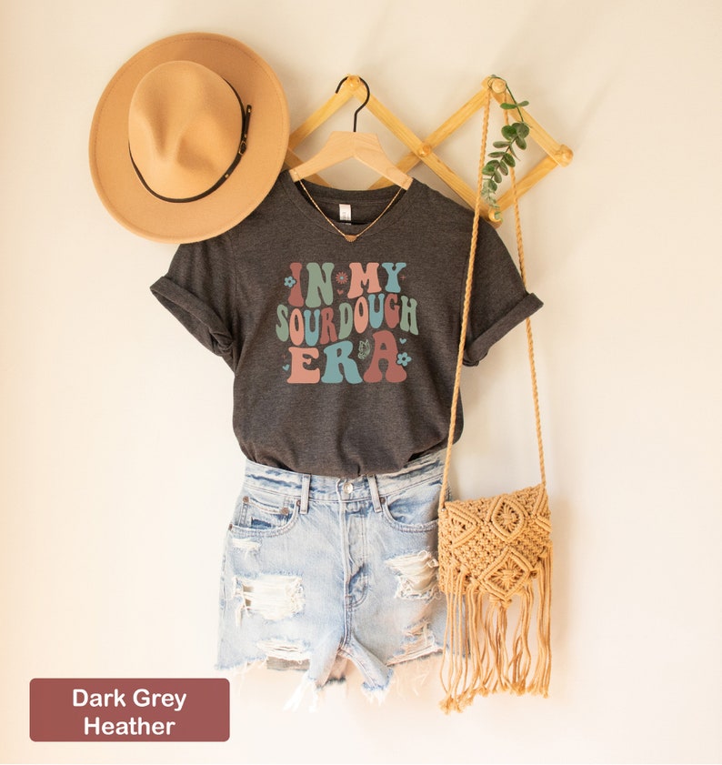 May include: Dark grey heather t-shirt with a retro-style graphic that says "In My Sourdough Era" with flowers and hearts. The shirt is hanging on a wooden hanger with a straw hat and a woven bag.