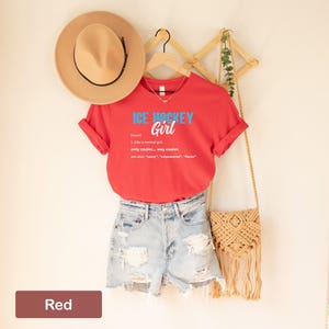 Op de afbeelding: Rood T-shirt met de tekst "ICE HOCKEY Girl" en een definitie van de term. Het shirt hangt aan een houten rek met een strohoed en een stro tas.