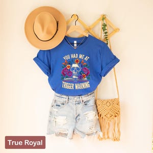 Puede incluir: Una camiseta azul con un gr&aacute;fico de calavera y flores y el texto "You had me at trigger warning".