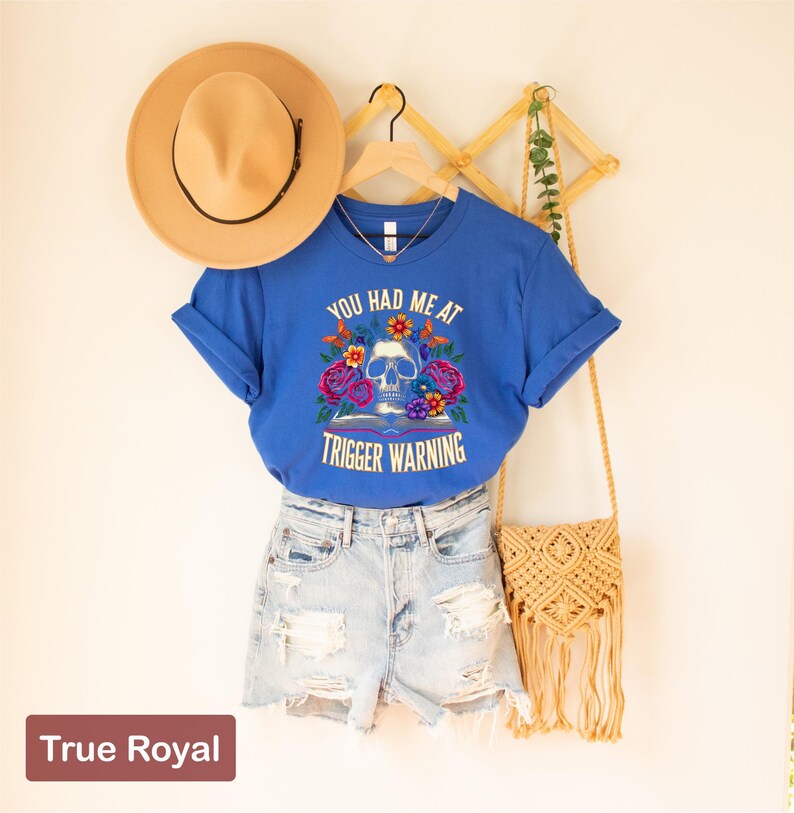 Puede incluir: Una camiseta azul con un gr&aacute;fico de calavera y flores y el texto "You had me at trigger warning".