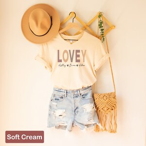 May include: A soft cream colored t-shirt with the word "LOVEY" printed in a gradient of brown, pink, and purple. The names "Rigley", "Brian", and "Adler" are printed below the word "LOVEY" with a heart symbol between each name.