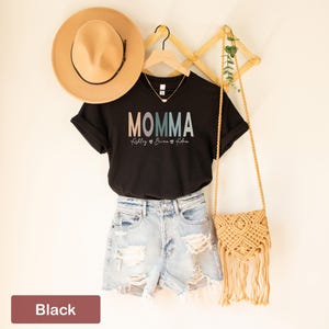 May include: Black t-shirt with the text "MOMMA" in rainbow colors and the names "Riley, Brianna, & Aiden" written below. The shirt is displayed on a hanger with a straw hat and a woven bag.