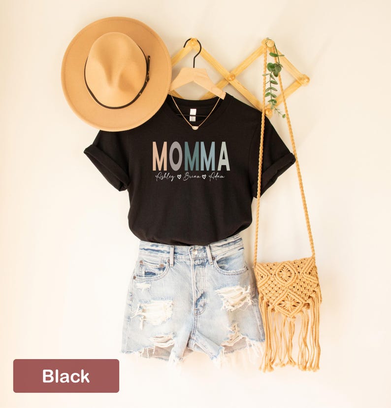 May include: Black t-shirt with the text "MOMMA" in rainbow colors and the names "Riley, Brianna, & Aiden" written below. The shirt is displayed on a hanger with a straw hat and a woven bag.
