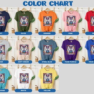 May include: Colour chart displaying various t-shirt colours featuring a colourful skull graphic and the text "Dia de los Muertos". The colours include navy, olive, orange, pink, red, silver, soft cream, steel blue, team purple, true royal, turquoise, white, and yellow.