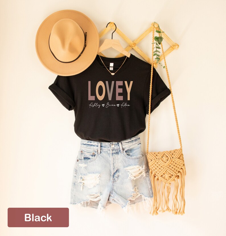 May include: Black t-shirt with the word "LOVEY" in a gradient of pink and brown. The names "Ashley", "Brian", and "Alena" are written below the word "LOVEY" with a heart symbol between each name.
