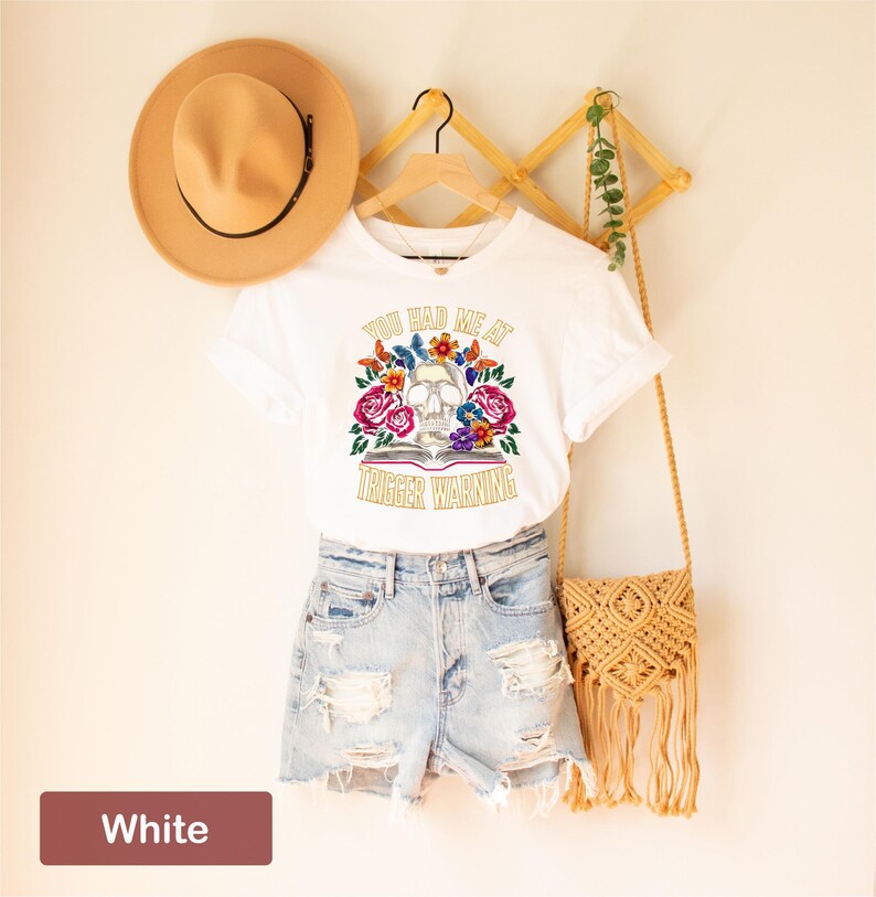 Puede incluir: Camiseta blanca con un gr&aacute;fico de calavera y flores y el texto "You had me at trigger warning".
