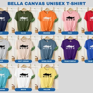 May include: A Bella Canvas unisex t-shirt with a graphic of a fish and the word "MEOW" printed on it. The shirt is available in 12 colors: navy, olive, orange, pink, red, silver, soft cream, steel blue, team purple, true royal, turquoise, white, and yellow.
