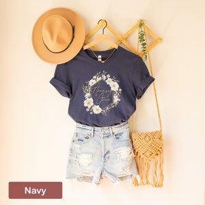 May include: Navy blue t-shirt featuring a floral wreath design and the text "Amazing Grace" in gold lettering.