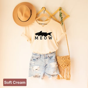 May include: A soft cream colored t-shirt with a black catfish silhouette and the word "MEOW" printed on the front. The shirt is hanging on a wooden hanger.