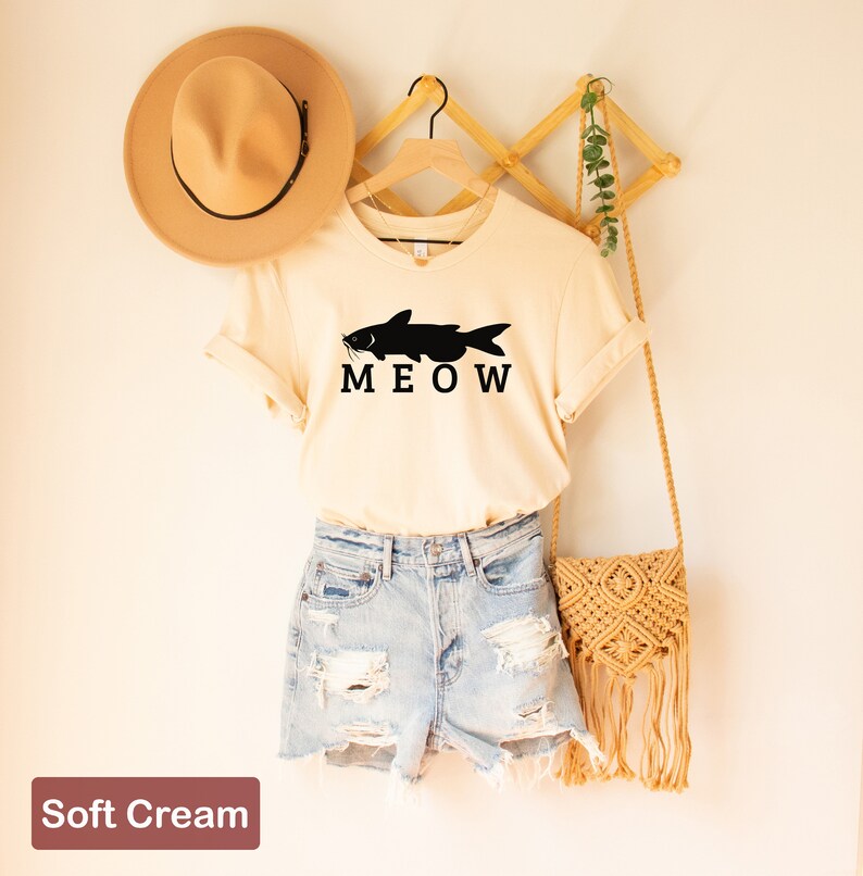 May include: A soft cream colored t-shirt with a black catfish silhouette and the word "MEOW" printed on the front. The shirt is hanging on a wooden hanger.