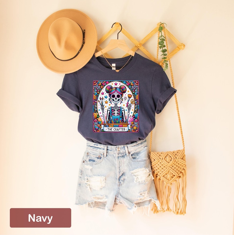 May include: A navy blue t-shirt featuring a colourful illustration of a skeleton wearing a blue overall and holding paintbrushes. The illustration is surrounded by a colourful border and the text "THE CRAFTERS" is at the bottom.