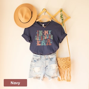 May include: A navy blue t-shirt with the text "In My Sourdough Era" in a retro font with colorful accents. The shirt is hanging on a wooden rack with a brown hat and a straw bag.