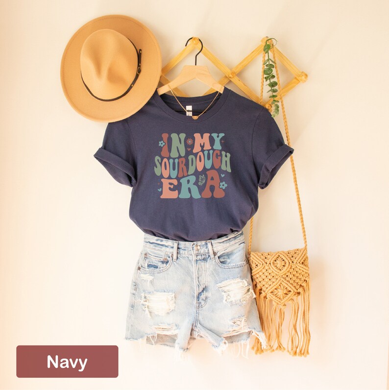 May include: A navy blue t-shirt with the text "In My Sourdough Era" in a retro font with colorful accents. The shirt is hanging on a wooden rack with a brown hat and a straw bag.