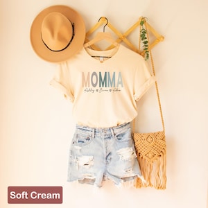 May include: A soft cream colored t-shirt with the word "MOMMA" in rainbow colors and the names "Lilly", "Brian", and "Adam" written underneath. The shirt is hanging on a wooden hanger with a straw hat and a straw bag.