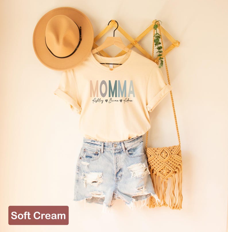 May include: A soft cream colored t-shirt with the word "MOMMA" in rainbow colors and the names "Lilly", "Brian", and "Adam" written underneath. The shirt is hanging on a wooden hanger with a straw hat and a straw bag.
