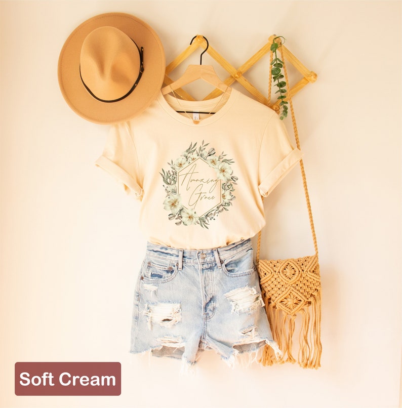 May include: A soft cream t-shirt featuring a floral design and the text "Amazing Grace" in gold lettering. The shirt is hanging on a wooden hanger and is styled with denim shorts and a straw bag.