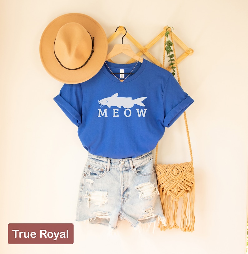 May include: A blue t-shirt with a white catfish graphic and the word "MEOW" printed on it. The shirt is hanging on a wooden hanger and is styled with a brown hat, denim shorts, and a straw bag.