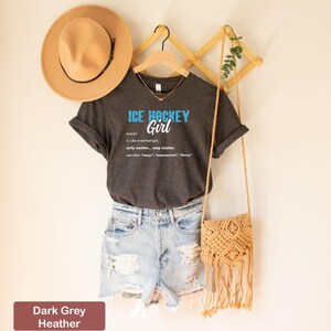 Op de afbeelding: Donkergrijs heather V-hals T-shirt met de tekst "ICE HOCKEY Girl" in blauw en wit. De tekst is gedefinieerd als "(zelfstandig naamwoord) 1. Net als een normaal meisje, alleen cooler... veel cooler. zie ook: "sassy", "empowered", "fierce""