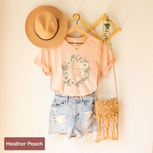 May include: A peach coloured t-shirt with a floral wreath design and the text "Amazing Grace" in gold lettering. The shirt is on a hanger and is styled with a straw hat and a crocheted bag.