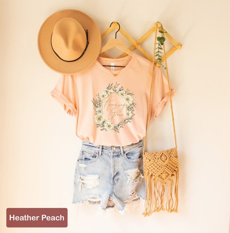 May include: A peach coloured t-shirt with a floral wreath design and the text "Amazing Grace" in gold lettering. The shirt is on a hanger and is styled with a straw hat and a crocheted bag.