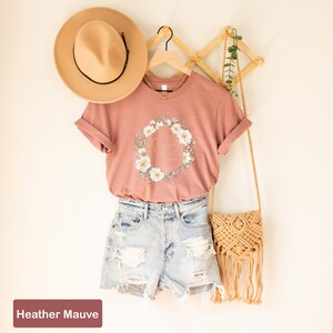 May include: A heather mauve t-shirt featuring a floral wreath and the text "Good Vibes Only" in gold lettering. The shirt is on a hanger and is styled with denim shorts and a straw bag.