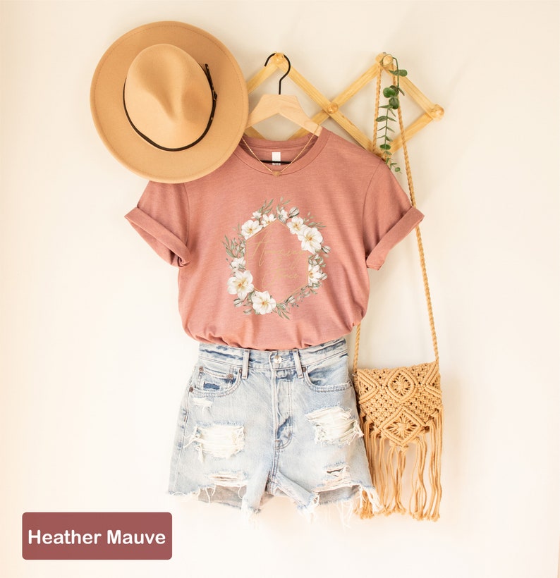 May include: A heather mauve t-shirt featuring a floral wreath and the text "Good Vibes Only" in gold lettering. The shirt is on a hanger and is styled with denim shorts and a straw bag.