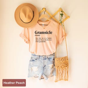 May include: A pink t-shirt with the text "Gramsicle (noun) One who sits at a hockey rink and freezes for the love of her grandchild." The shirt is hanging on a wooden hanger with a brown hat and a woven bag.