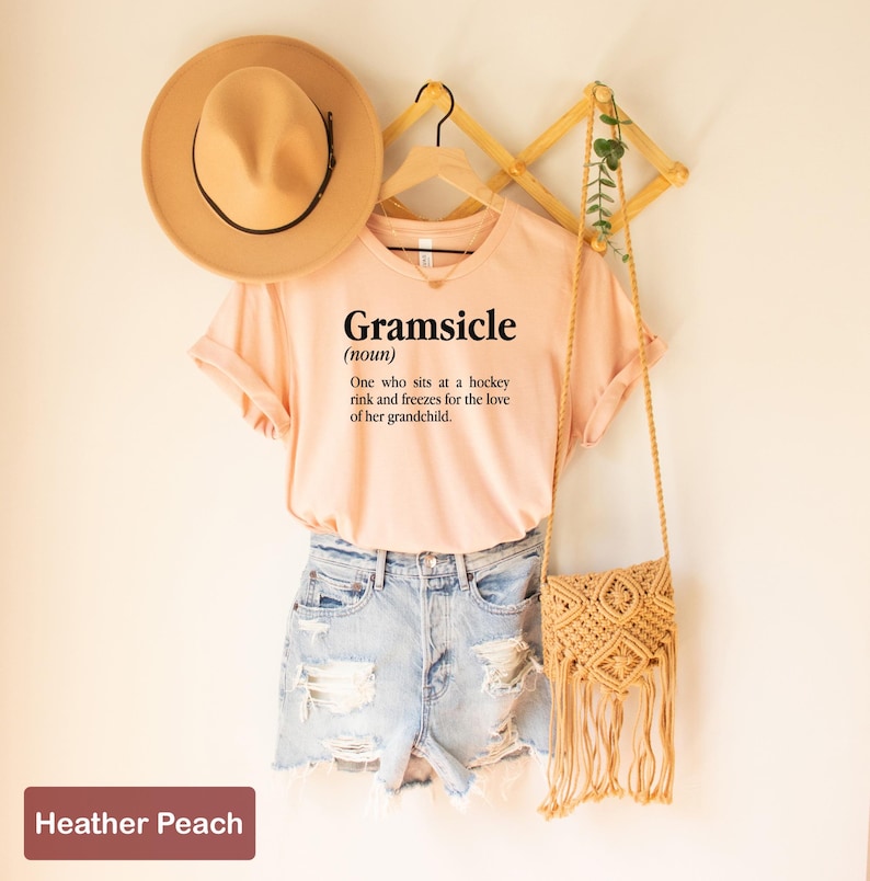 May include: A pink t-shirt with the text "Gramsicle (noun) One who sits at a hockey rink and freezes for the love of her grandchild." The shirt is hanging on a wooden hanger with a brown hat and a woven bag.