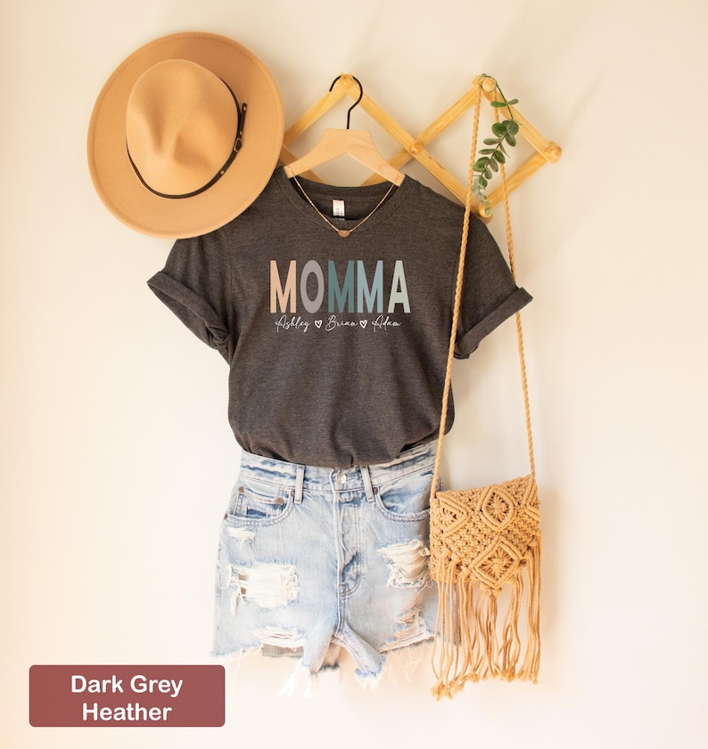 May include: Dark grey heather V-neck t-shirt with the word "MOMMA" in colorful block letters and the names "Ashley", "Brian", and "Adam" written in a script font below. The shirt is hanging on a wooden hanger with a straw hat and a woven bag.